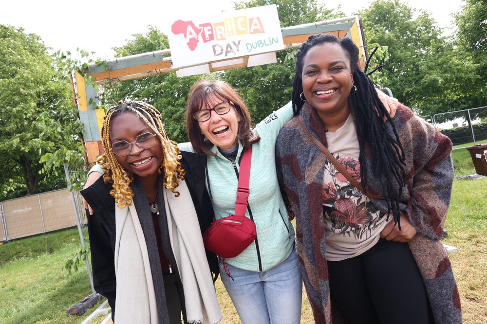 three women with their arms around each other in front of a wooden sign with Africa Day