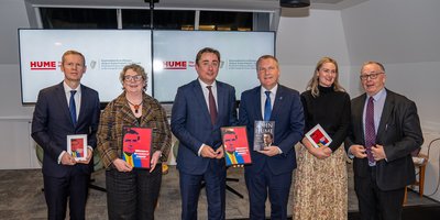 Don Sexton from the Irish Permanent Representation, Ambassador Aingeal O'Donoghue, John Hume Jr., Commissioner Michael McGrath, Shona Murray and Séamus Boland all pose together at the front of the stage. They hold framed red posters of John Hume.