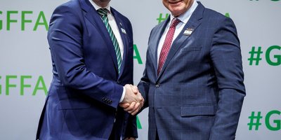 Two men in suits stand shaking hands in front of a white backdrop, which has '#GFFA' printed in green multiple times