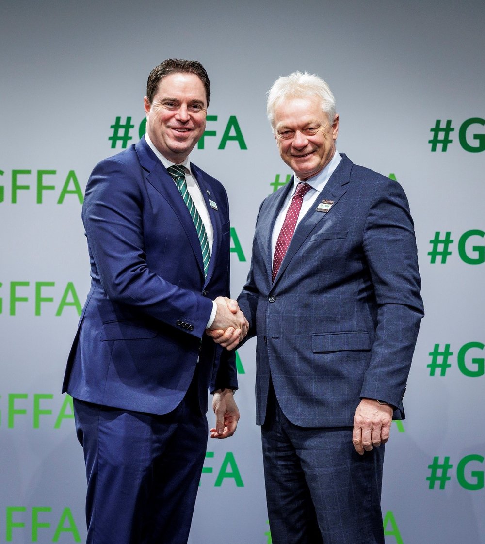 Two men in suits stand shaking hands in front of a white backdrop, which has '#GFFA' printed in green multiple times