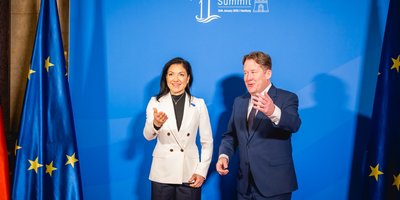 Germany's Minister Katharina Reiche greets Ireland's Minister Darragh O'Brien, both with outstreteched arms. Reiche is wearing a white blazer and black top, O'Brien is wearing a navy suit and white shirt. They stand in front of a blue backdrop.