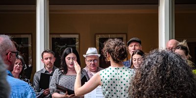 Choir singing with female conductor standing in front of them with her back to the camera.