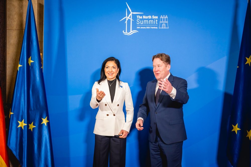 Germany's Minister Katharina Reiche greets Ireland's Minister Darragh O'Brien, both with outstreteched arms. Reiche is wearing a white blazer and black top, O'Brien is wearing a navy suit and white shirt. They stand in front of a blue backdrop.