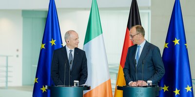 Taoiseach Micheál Martin and Chancellor Friedrich Merz stand at a podium each speaking at a press conference.