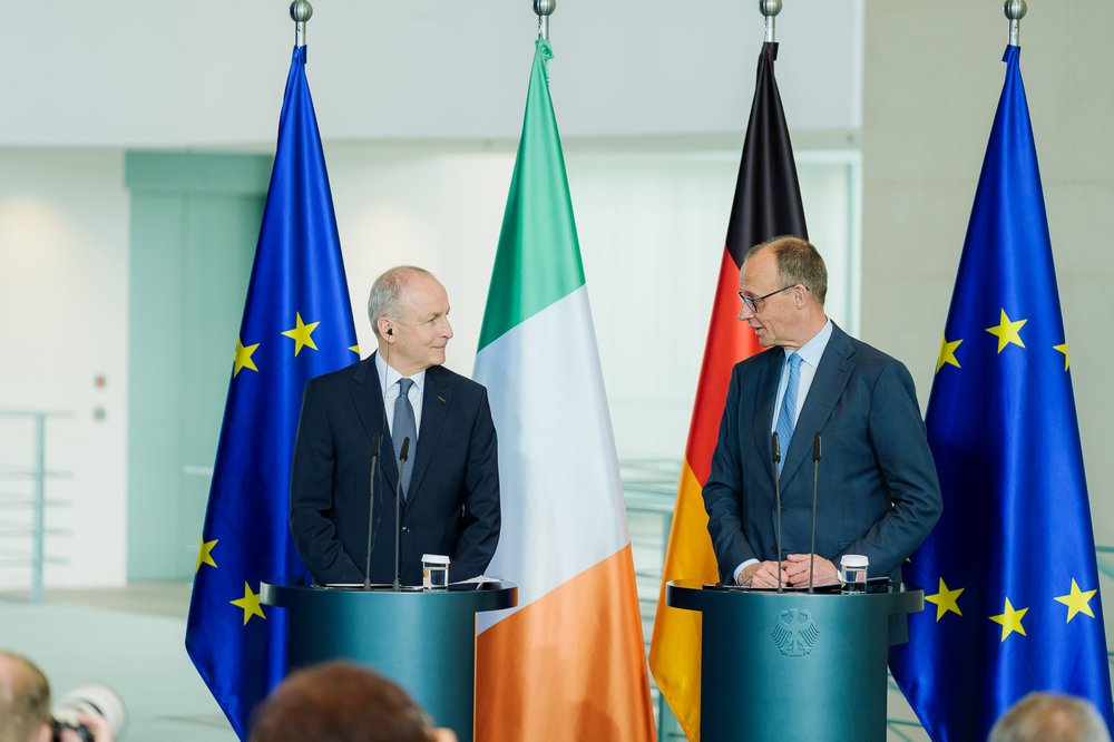 Taoiseach Micheál Martin and Chancellor Friedrich Merz stand at a podium each speaking at a press conference.