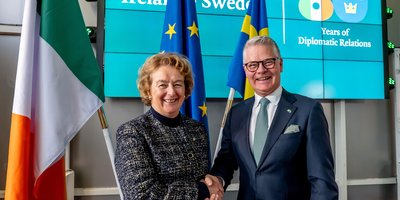 Ambassador Barbara Jones stands with Honorary Consul to Ireland in Gothenburg Niclas Mårtensson smiling and shaking hands. They stand in front of an Irish, European, and Swedish flag, and a screen displaying a logo symbolising 80 years of Diplomatic ties between Ireland and Sweden.