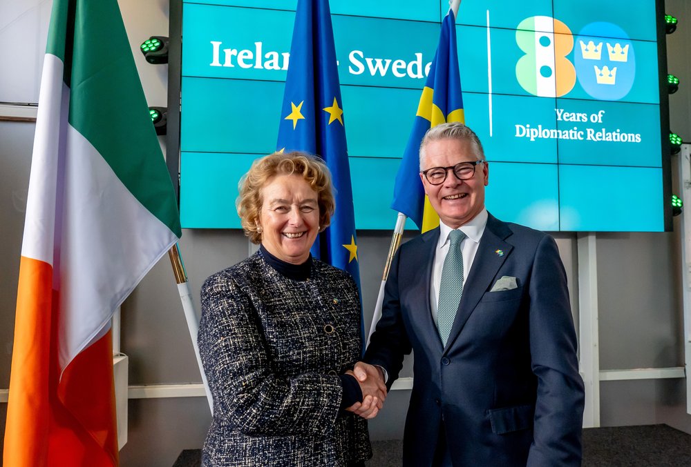 Ambassador Barbara Jones shakes hand with Honorary Consul Niclas Mårtensson. They stand in front of the Irish, EU, and Swedish flags, and a screen displaying a logo symbolising 80 years of diplomatic ties between Ireland and Sweden.