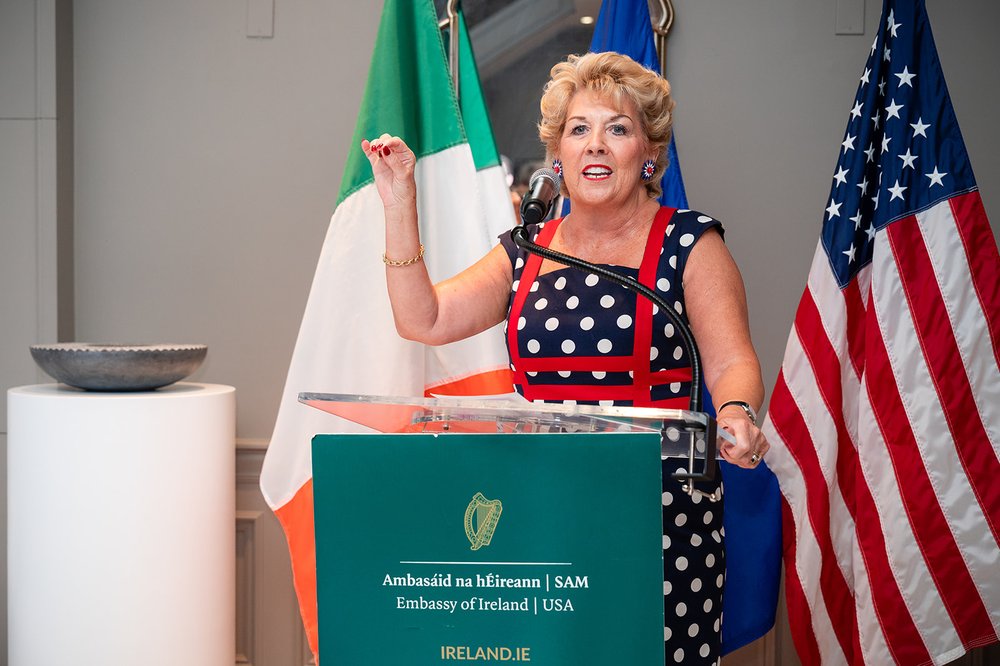 Ambassador Geraldine Byrne Nason speaks at podium in front of Irish, EU, and American flags in a red and blue polka dot desk at reception marking 250 years of shared history between Ireland and America.