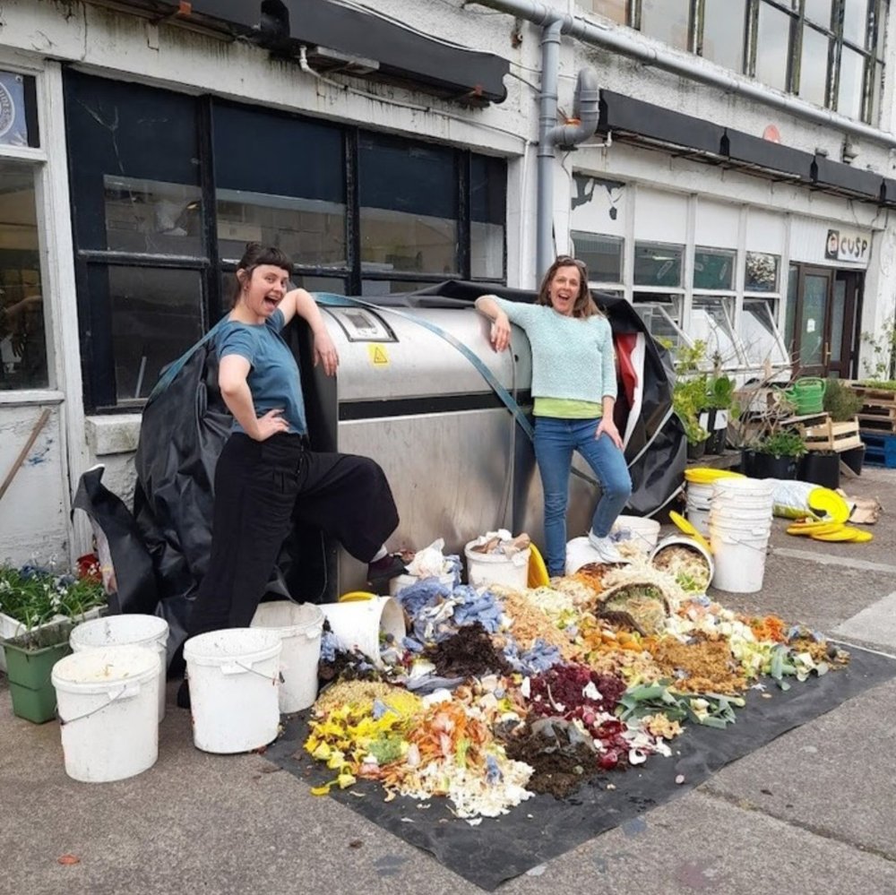 Two women pose in front of the CUSP biodigester with food waste scraps and buckets at their feet