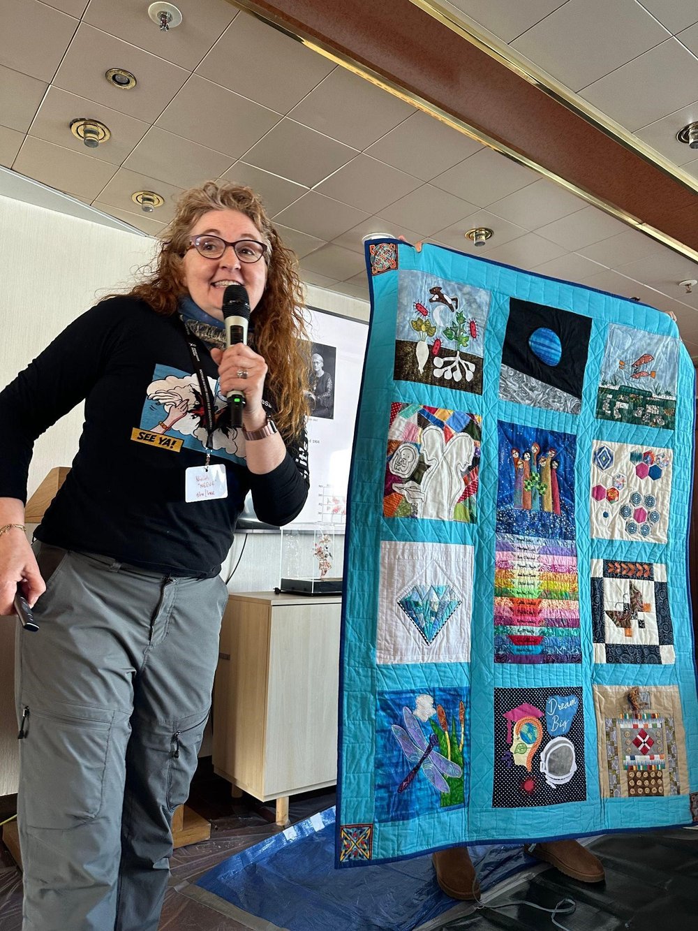 Dr Niamh Shaw speaks into a microphone next to a twelve-panel quilt featuring Irish women in science, which is displayed vertically