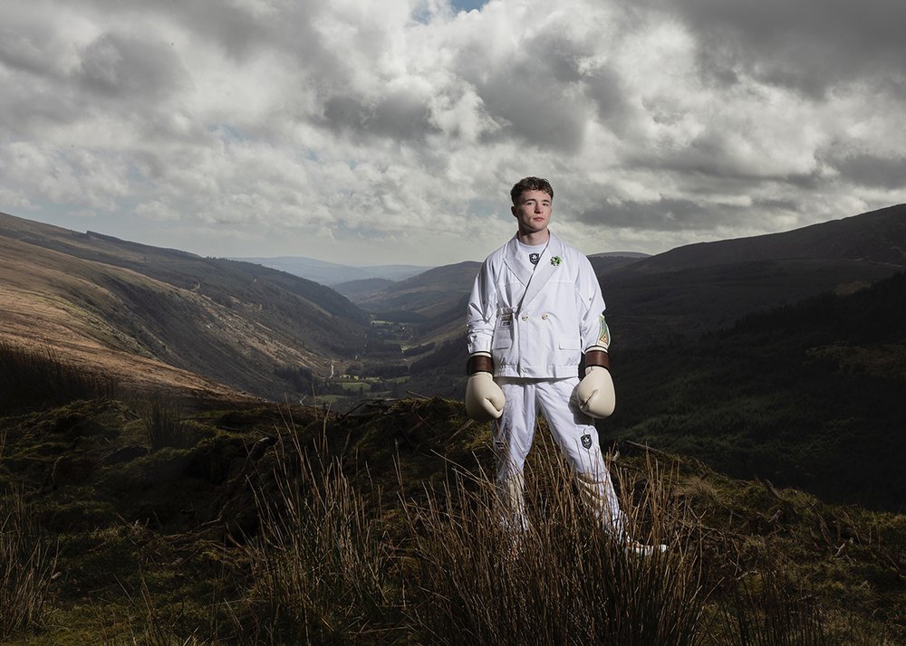 A man dressed in the white Irish Olympic uniform and boxing gloves standing in a mountainous landscape.