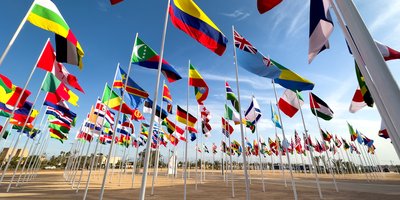 Flags of several countries flying on tall flagpoles, as seen from the ground.