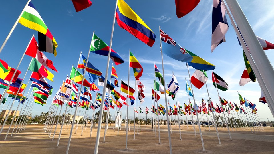 Flags of several countries flying on tall flagpoles, as seen from the ground.