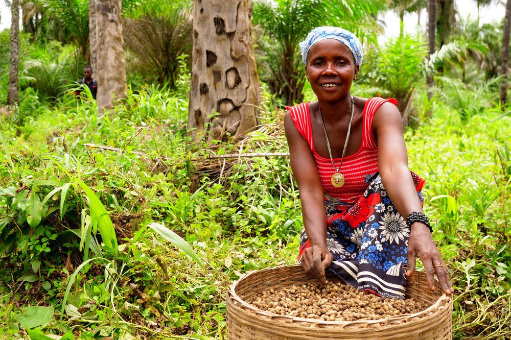 a woman surrounded by greenery holding a basket of grain and wearing colourful clothes