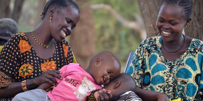 Two women smile as they watch two toddlers lean their heads together, as if one child is whispering in the ear of the other.