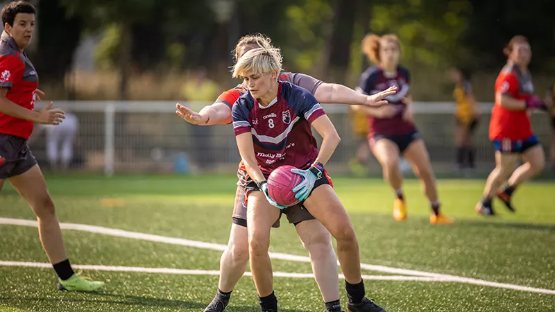 one woman holding a ball on a pitch with a player behind her reaching out either side