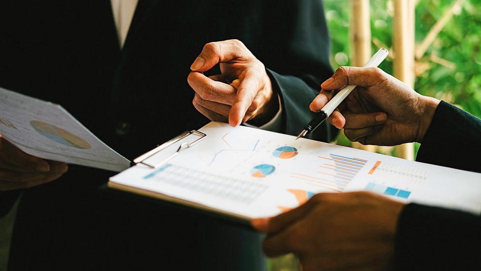 Close-up of a page on a clipboard with charts and graphs. The hand of the person holding the clipboard is hovering over the page with a pen while another person's hand can be seen pointing at a part of the page.