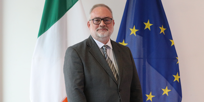 Ambassador Kevin Conmy standing in front of the Irish and EU flags.