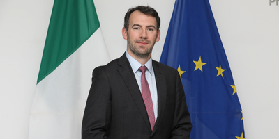 A man wearing a suit standing in front of the Irish and EU flags.