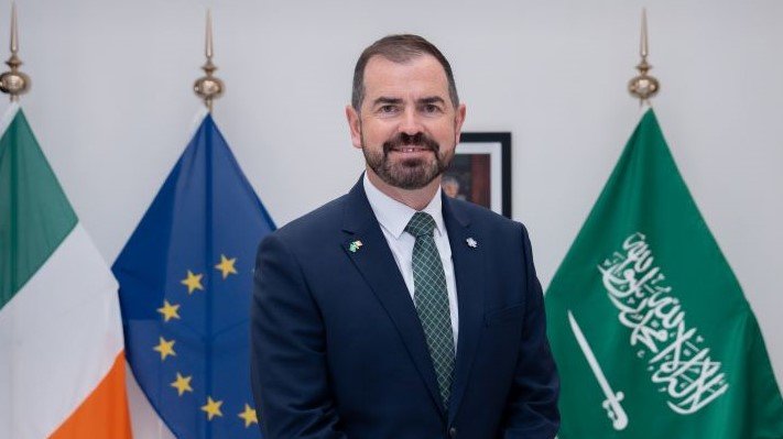 The Ambassador in front of the Irish, EU and Saudi flag.