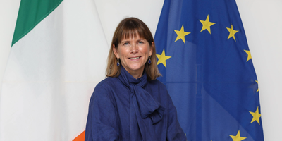 Headshot of Alison Milton standing in front of the Irish and EU flags