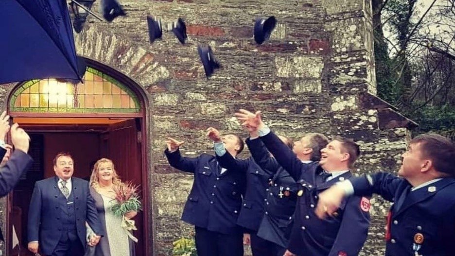 People throwing their hats in the air as newlyweds walk out of a house