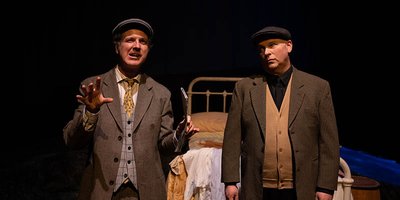 two men in costume from 100 years ago with old bed behind them on stage