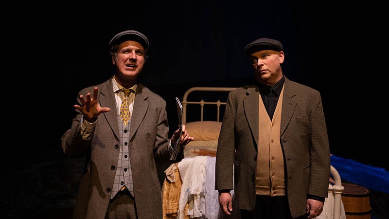 two men on stage dressed in costume from 100 years ago with an old bedframe behind them