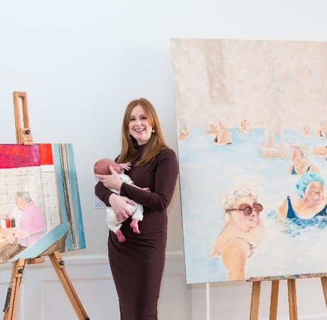 Artist holding her baby and standing beside her paintings