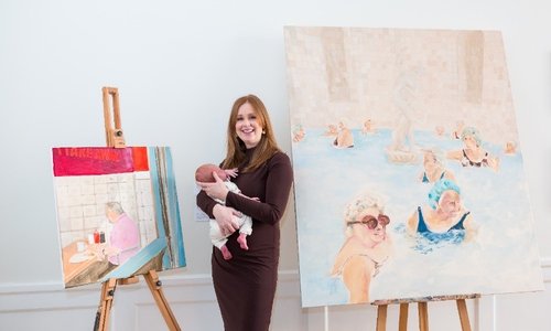 Artist holding her baby and standing beside her paintings