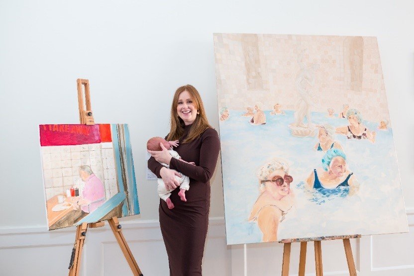 Artist holding her baby and standing beside her paintings