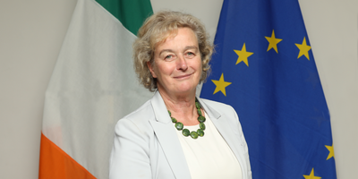 Headshot of a blonde woman with short hair standing in front of the Irish and EU flags.