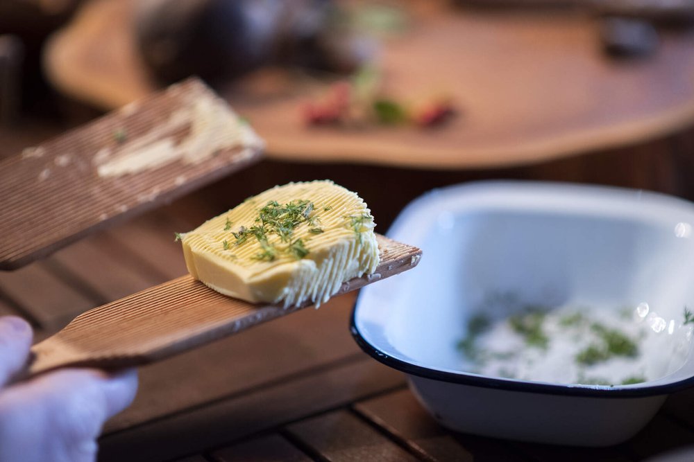 a pat of butter on a wooden butter paddle, it is garnished with herbs