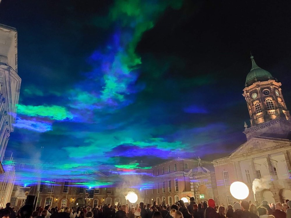 A crowd is gathered underneath a smoke and coloured laser canopy at night at Dublin Castle.