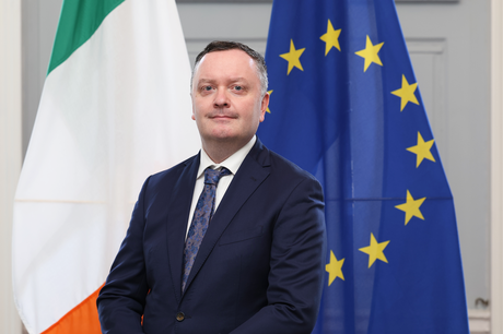 Headshot of a man with short black hair and dark navy jacket with tie standing in front of the Irish and EU flags.
