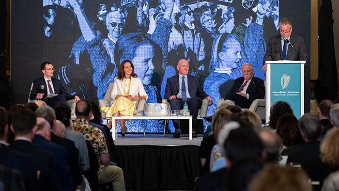 four people sitting on stage for a panel, with a screen of crowd photography behind them