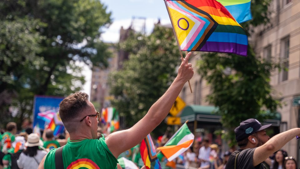 A man in a green tshirt with a rainbow on the back raising the Progress Pride Flag during the New York City Pride March