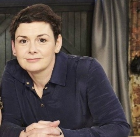 Caitrona Carney is wearing a navy shirt with her hands clasped in front of her with a slightly smiling expression