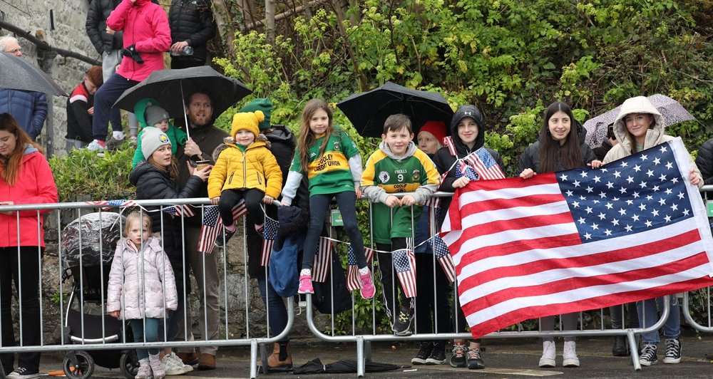 crowds waiting behind barrier on side of road with American flag