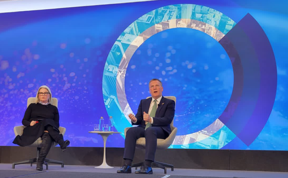 a woman and a man sitting on a stage with a table between them and a blue screen behind them
