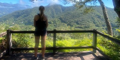 A woman with her back to the camera looks at a sun-lit valley and mountain range in the distance.