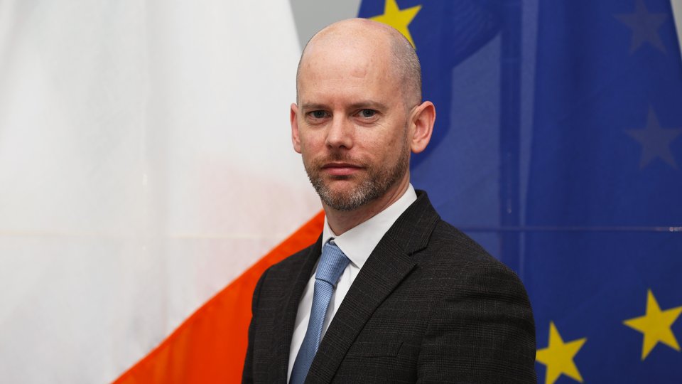 Consul General Cillian O'Kelly in front of Irish and EU flags.