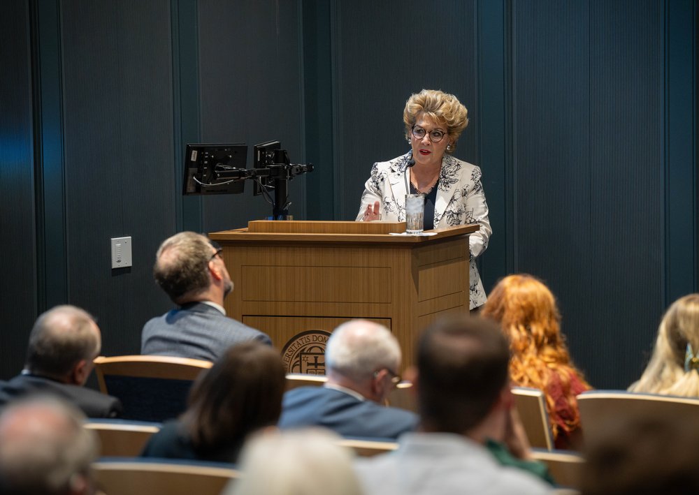 Ambassador Byrne Nason speaks to audience at podium in blue auditorium.