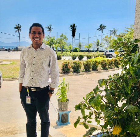 A man stands in front of a sun-filled square lined with trees and plants.