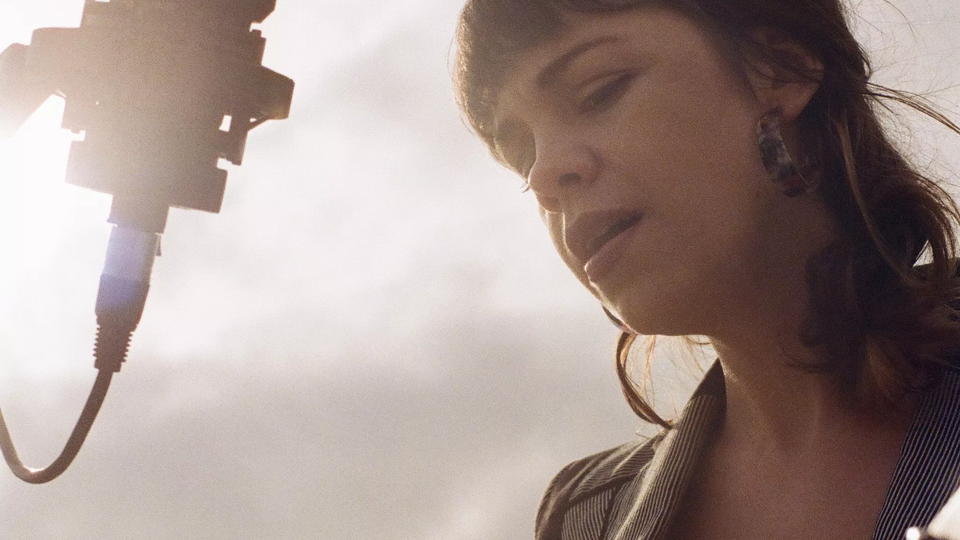 one woman playing guitar and singing into a microphone outside with sun shining