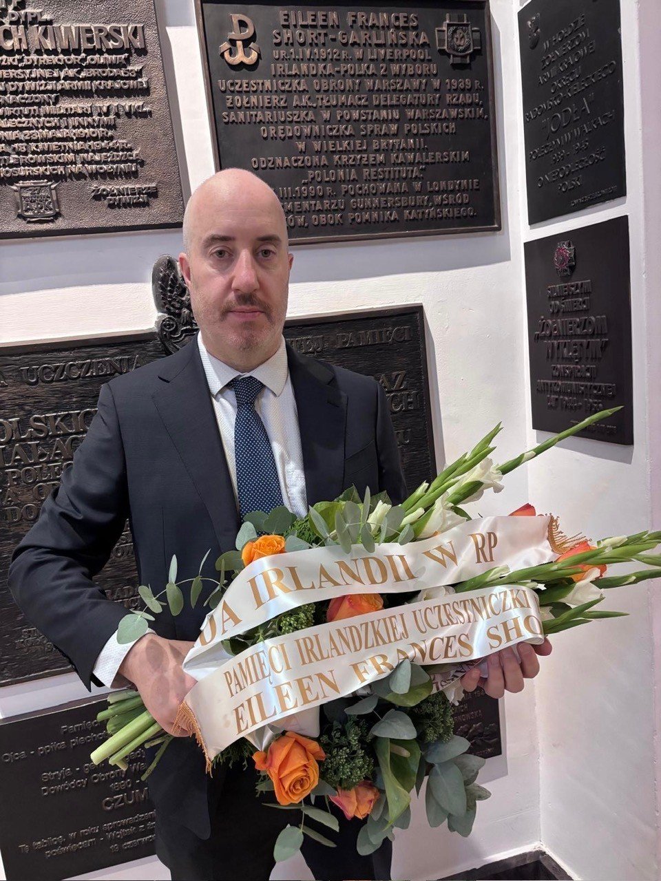 Deputy HOM, Brendan Farrell holding flowers, in front of a nameplate commemorating Eileen Frances Short-Garlinska