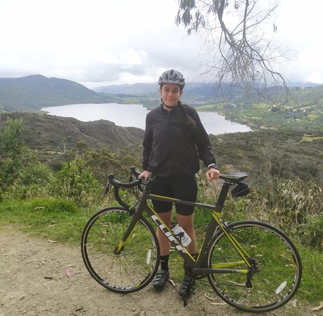 A woman wearing a helmet stands behind a bike. In the background is a lake and green mountains.