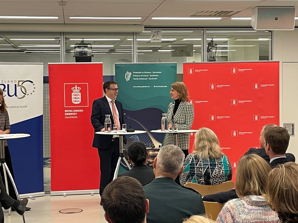 Two members of the panel standing on stage in front of banners for the Swedish and Irish Embassies.