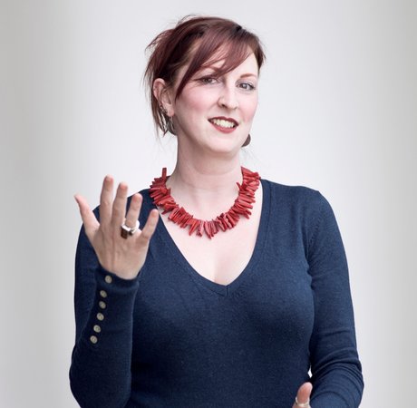 Emma is wearing a bright statement necklace and has one hand raised as she smiles softly