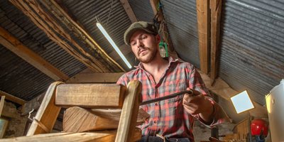 Eoin Reardon in a rustic stone-walled workshop, he is smiling leaning against a chair that is on a workbench laiden with sawdust. Eoin is holding a mallet with a wooden sign reading "Eoin Reardon Woodcraft"